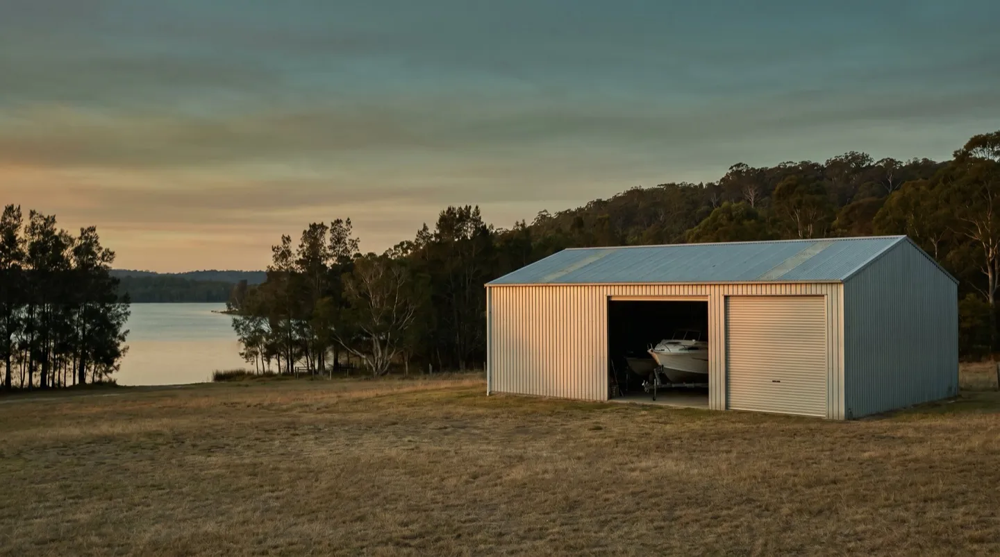 A large galvanised farm shed with high roller doors on an acreage block, a boat on a trailer inside, bushland ridge behind