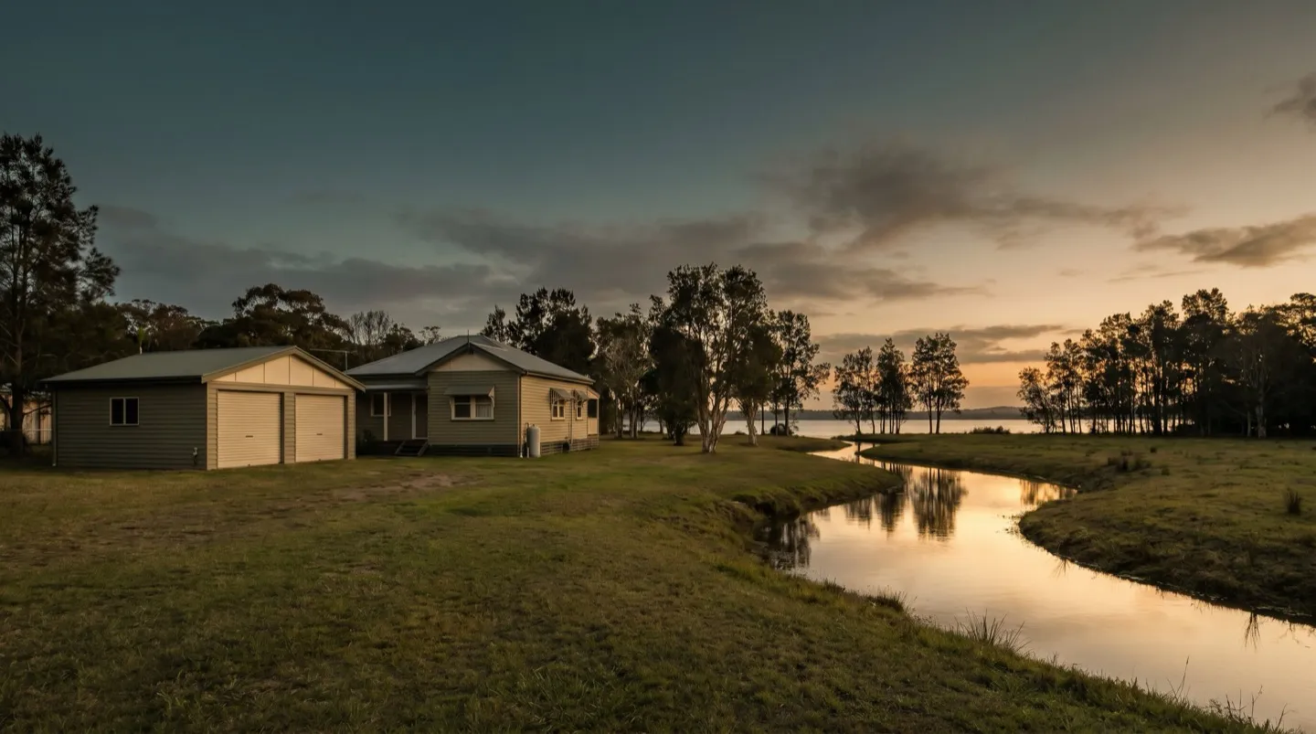 A rural-residential house and separate double garage with roller doors beside a still creek at dusk